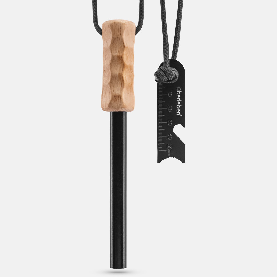 The Kräftig Grip Fire Starter by Überleben features a black survival fire starter tool with a handcrafted hardwood handle, hanging from a black cord. Attached to the cord is a metal scraper with measurement markings and a reliable ferrocerium rod for efficient spark generation.