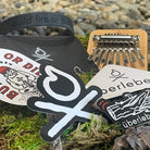 A Welcome Pack from Überleben displayed on a mossy surface, featuring a wristband with "build fire or die" text, several stickers showcasing graphics and the word "überleben," an instrument that looks like a kalimba, and a partially visible black object in the background. Pebbles can be seen in the distance.