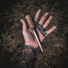 A tattooed hand holds the Überleben Kräftig Grip Fire Starter, featuring a handcrafted hardwood handle and a metal striker. The background is an outdoor setting with dried grass and sparse vegetation, suggesting a wilderness or camping environment.