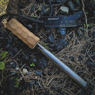 A fire starter with a handcrafted hardwood handle, named "Kräftig Grip" by Überleben, lies on the ground amid twigs and leaves. A black ruler labeled "überleben," measures from 0 to 50mm next to it. The Kräftig Grip includes a ferrocerium rod and features a black cord and multi-tool striker visible in the background.