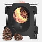 The Stöker | Stove by Überleben is a black, sleek wood stove with a circular opening displaying a bright, roaring fire inside. Two pinecones are placed in front of the stove, adding a natural touch. Crafted from stainless steel, this stove showcases a simple, minimalist design on a plain white background.