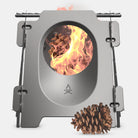 Close-up of the Überleben Stöker stove with a fire burning inside, fueled by leaves. A hand is adjusting a grill on top of the stainless steel stove. The image captures an outdoor setting with a blurred natural background, highlighting the compact metal structure of the Stöker stove.