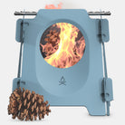 The Überleben Stöker is a portable, compact twig stove in blue, featuring a circular opening that emits flames and contains a pinecone inside. This stainless steel stove comes with foldable legs and has a pinecone lying beside it on the ground. It boasts a small logo with flame and water drop icons and is designed for use with organic fuel.