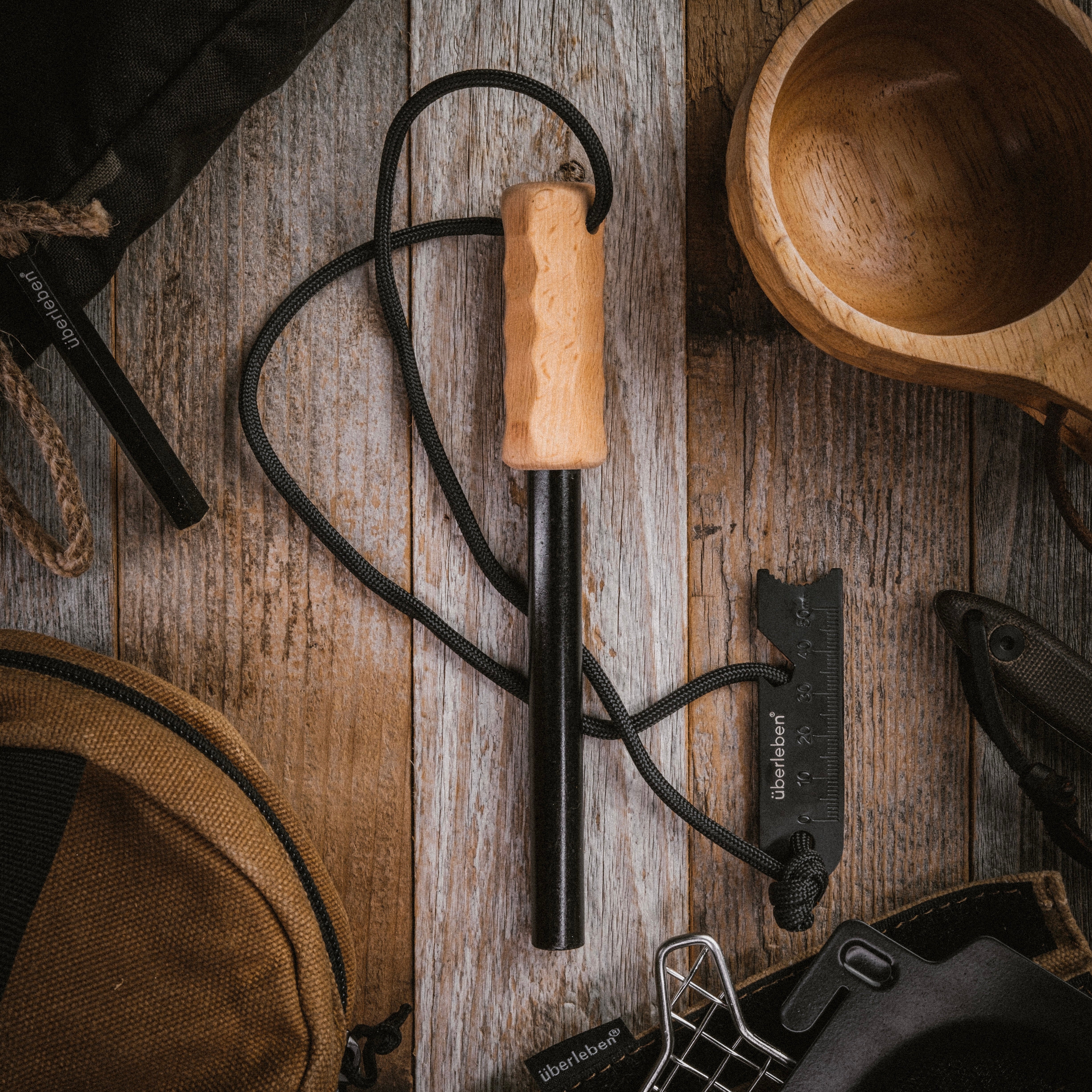 A handcrafted hardwood handle adorns the black stick of the Überleben Kräftig Grip Fire Starter, complemented by a wooden cup on a wood surface.