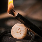 Close-up image of a candle wick being lit using the Überleben Kräftig Grip | Fire Starter. The candle’s lid, made of handcrafted hardwood, features an engraved heart and the letters "Xo." A black cord is partly visible, possibly part of the candle's packaging. The background is blurry, with muted colors.