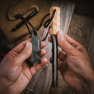 Close-up of hands holding the Kräftig Grip | Fire Starter by Überleben, a black multitool ruler equipped with a bottle opener and a handcrafted hardwood handle on the ferrocerium rod. The items are connected by black cords. The background includes a brown pouch, rope, and a wooden surface.