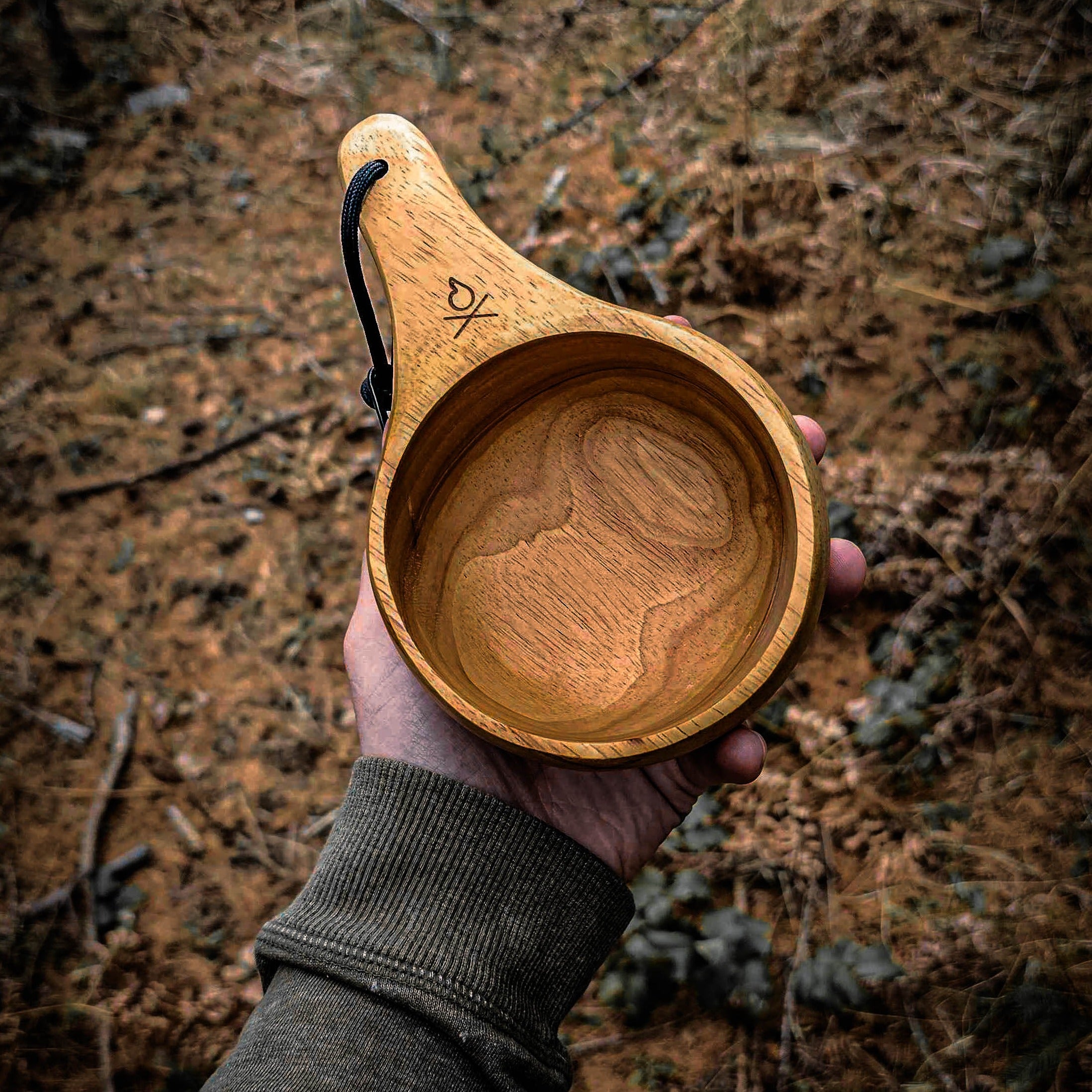 A hand holds the Lore Kuksa by Überleben, a handcrafted piece featuring a black paracord rope. Made from 100% natural hardwood, this kuksa boasts a smooth, polished surface and a carved fire icon on the handle. 