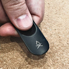 A close-up of a person's fingers holding a black metal striker with a small fire icon on the surface. The background is textured cork material, highlighting the sleek design of the modern Überleben Ambistriker, which is perfect for left handed or right handed people. all-groups