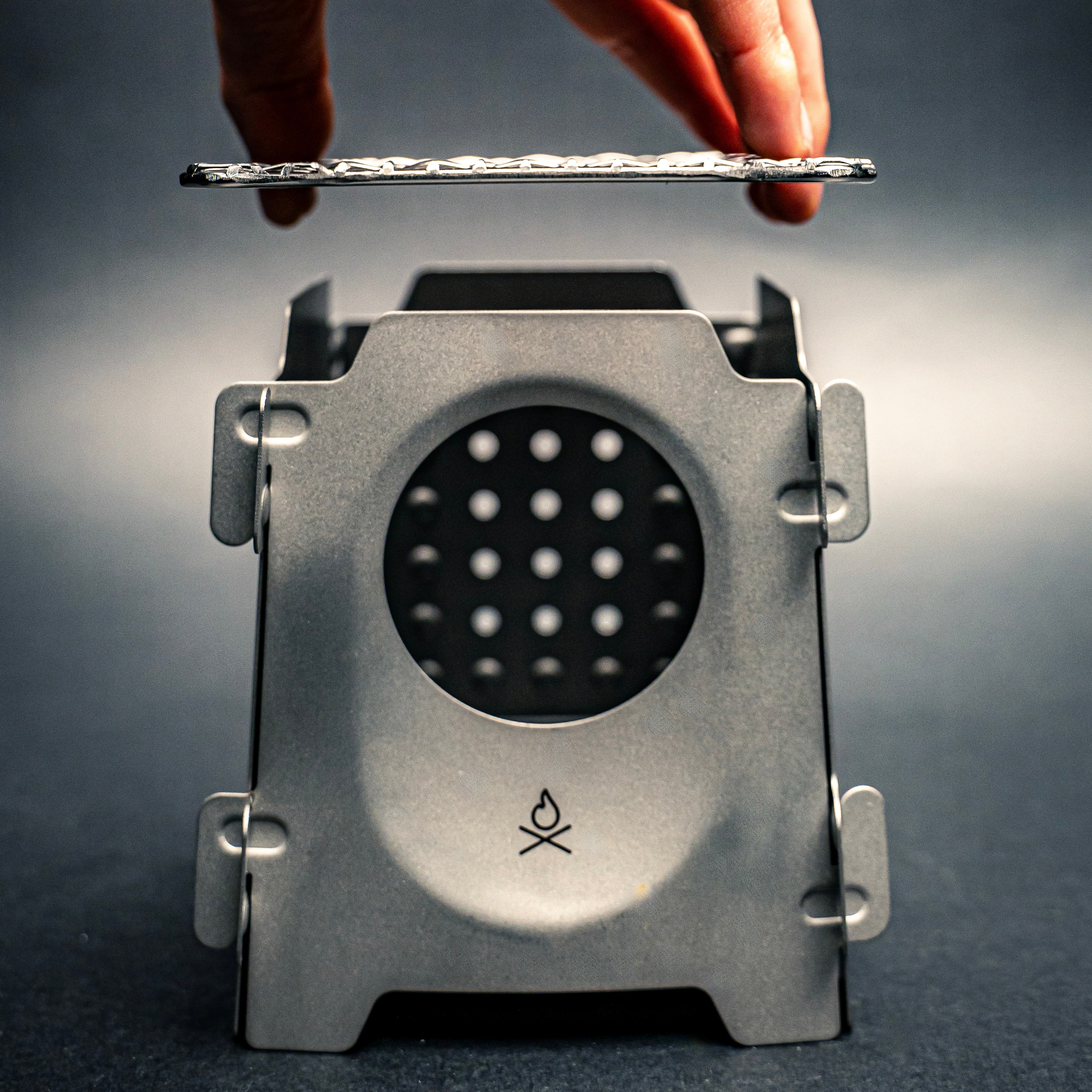 A close-up of the bushcraft Stöker Stove, a twig stove, by Überleben, showcasing its bead blasted surface. A hand is dropping a grate on top of the stove.