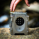 A hand is seen placing a small,  stainless steel twig stove with a circular opening, on a rocky surface. The Überleben Stöker Stove which uses organic fuel, features a simple fire icon on the front.