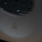 Close-up image of the Überleben Stöker stove, featuring perforated holes and the brand text "überleben" clearly visible. The foreground includes an out-of-focus grey element partially obstructing the view.