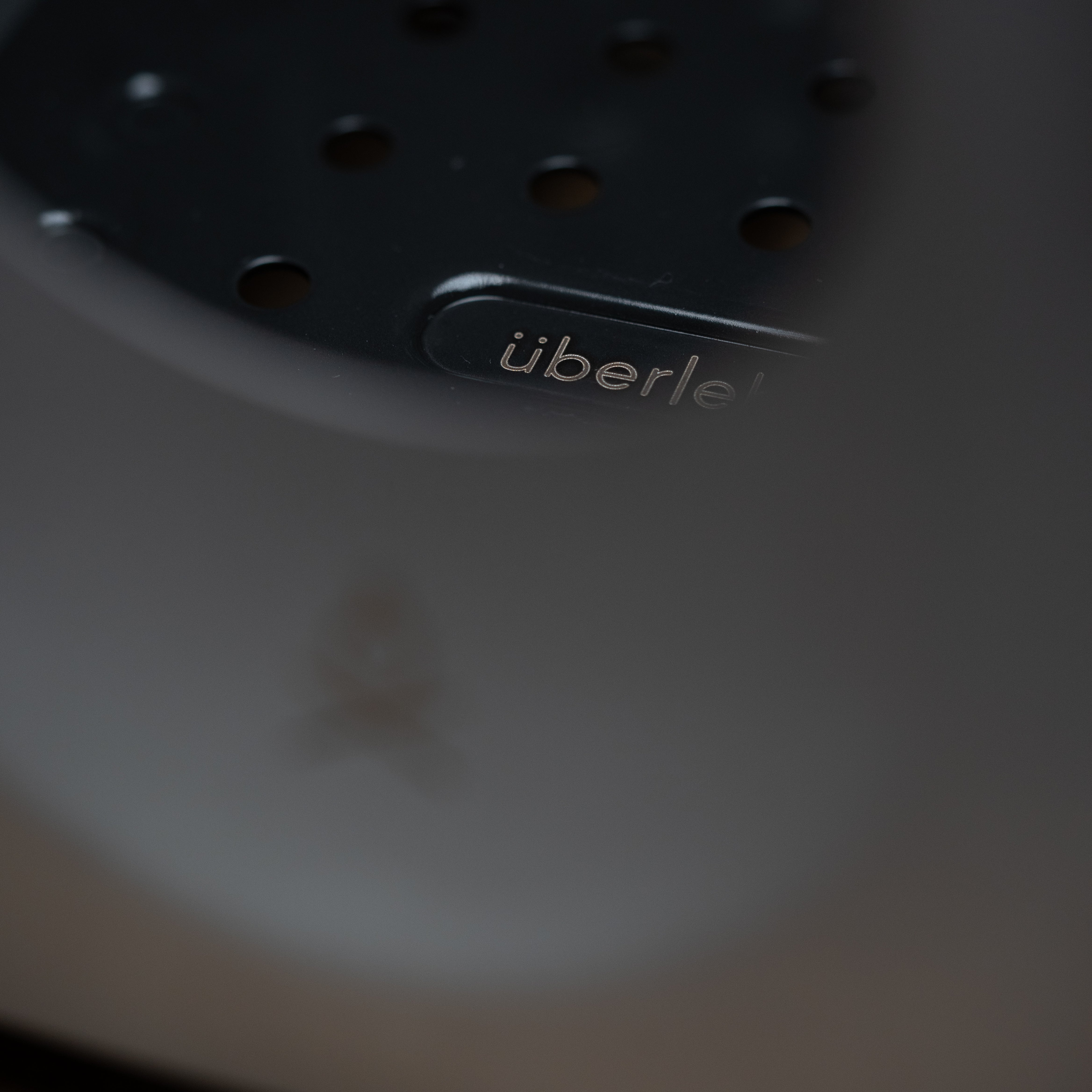 Close-up image of the Überleben Stöker stove, featuring perforated holes and the brand text "überleben" clearly visible. The foreground includes an out-of-focus grey element partially obstructing the view.