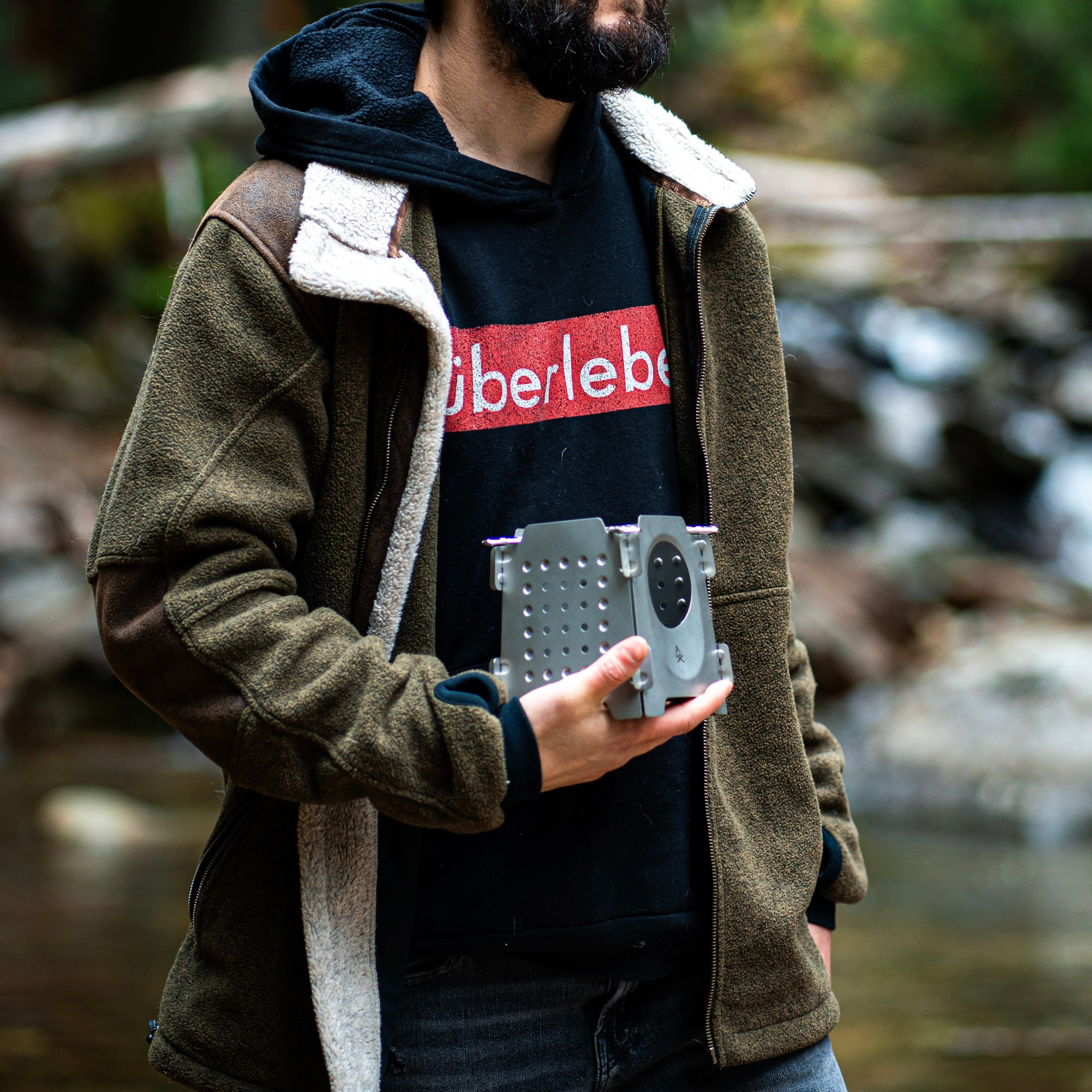 A person wearing a fleece jacket over a hoodie that features the word 'Überleben' stands outdoors, holding an Überleben Stöker stainless steel bushcraft twig stove. The background showcases a blurred natural setting with trees and a stream.