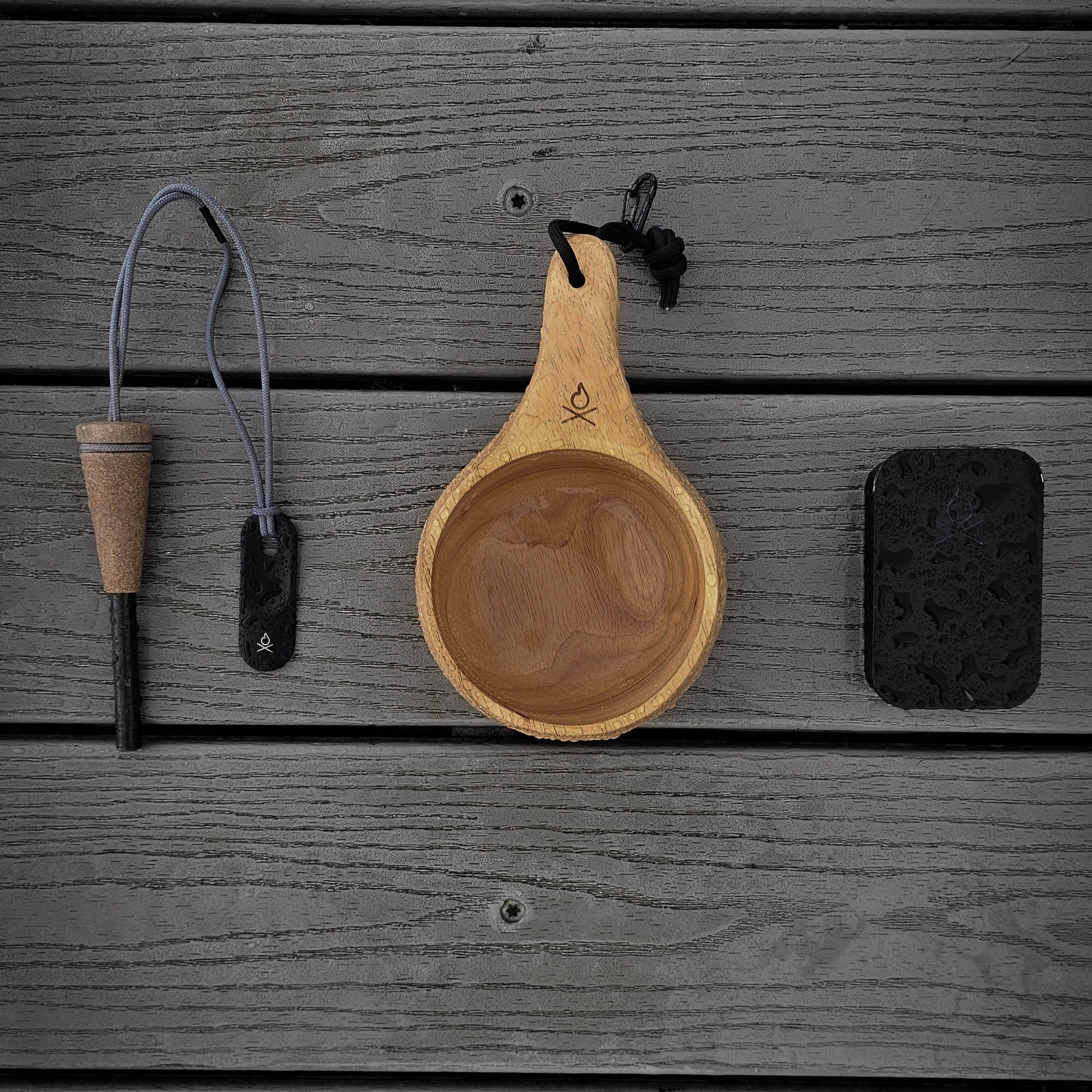 Flat lay of camping tools on a boat dock. From left to right: a Zünden fire starter with striker and wooden handle, the Lore Kuksa by Überleben made from 100% natural hardwood, and a small fire kit tin. Each item is arranged horizontally.