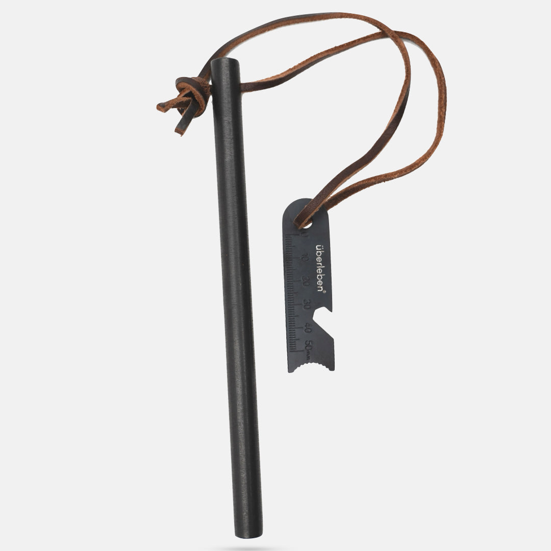 The Überleben Kräftig Fire Starter is a black ferro rod equipped with a multi-tool striker and attached via a brown leather cord. The striker features measuring marks and a bottle opener, making it ideal for outdoor and survival use.