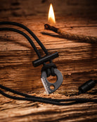 A black survival fire starter identified as the Überleben Leicht | Wearable Fire Starter, is displayed on a rustic wooden surface. In the background, a small flame burns on a jute rope. The fire starter includes 550 paracord for added versatility, and is great for bushcraft/survival situations