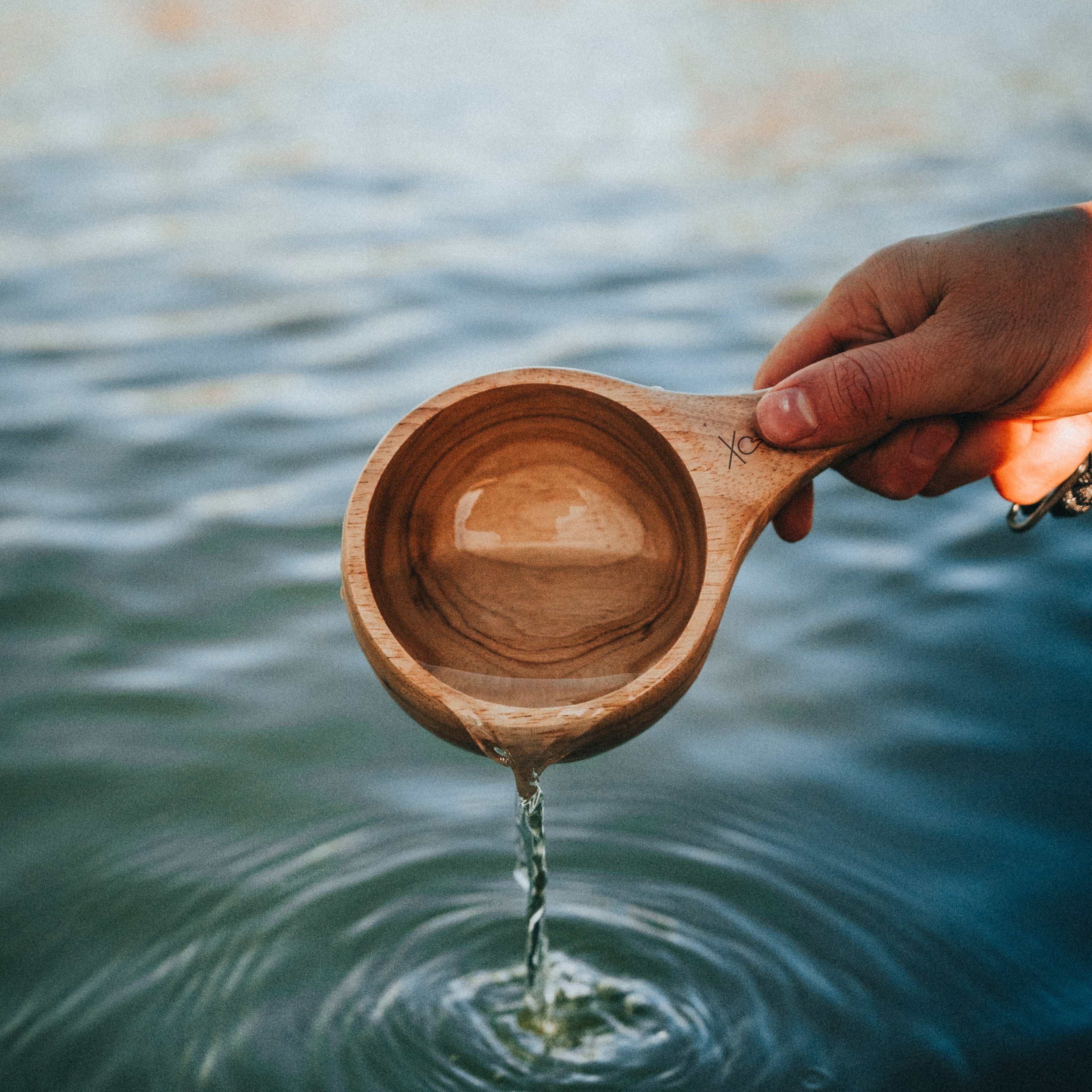 A hand holds the Lore Kuksa by Überleben, scooping water from a lake. This handcrafted kuksa, made from 100% natural hardwood, boasts a smooth, polished finish. 