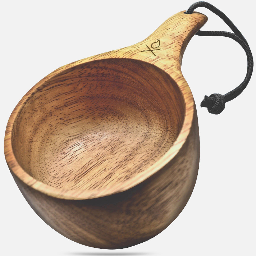 The Lore  Kuksa by Überleben is displayed, showcasing its smooth-finished wooden surface and rounded design. The handle has a fire icon engraved into it and includes black 550 paracord threaded through a hole for convenient hanging. 