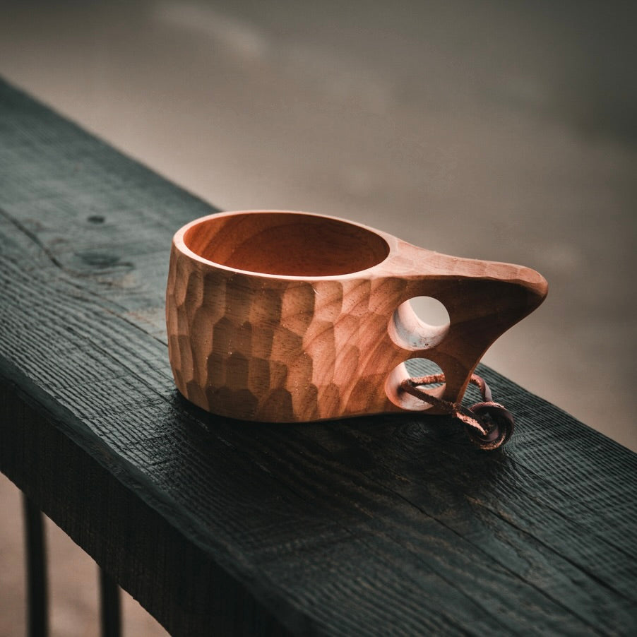 The Überleben Original 8oz. Kuksa, a handcrafted camp cup made from natural hardwood and featuring a carved design with a handle that has two holes, sitting on a porch railing.