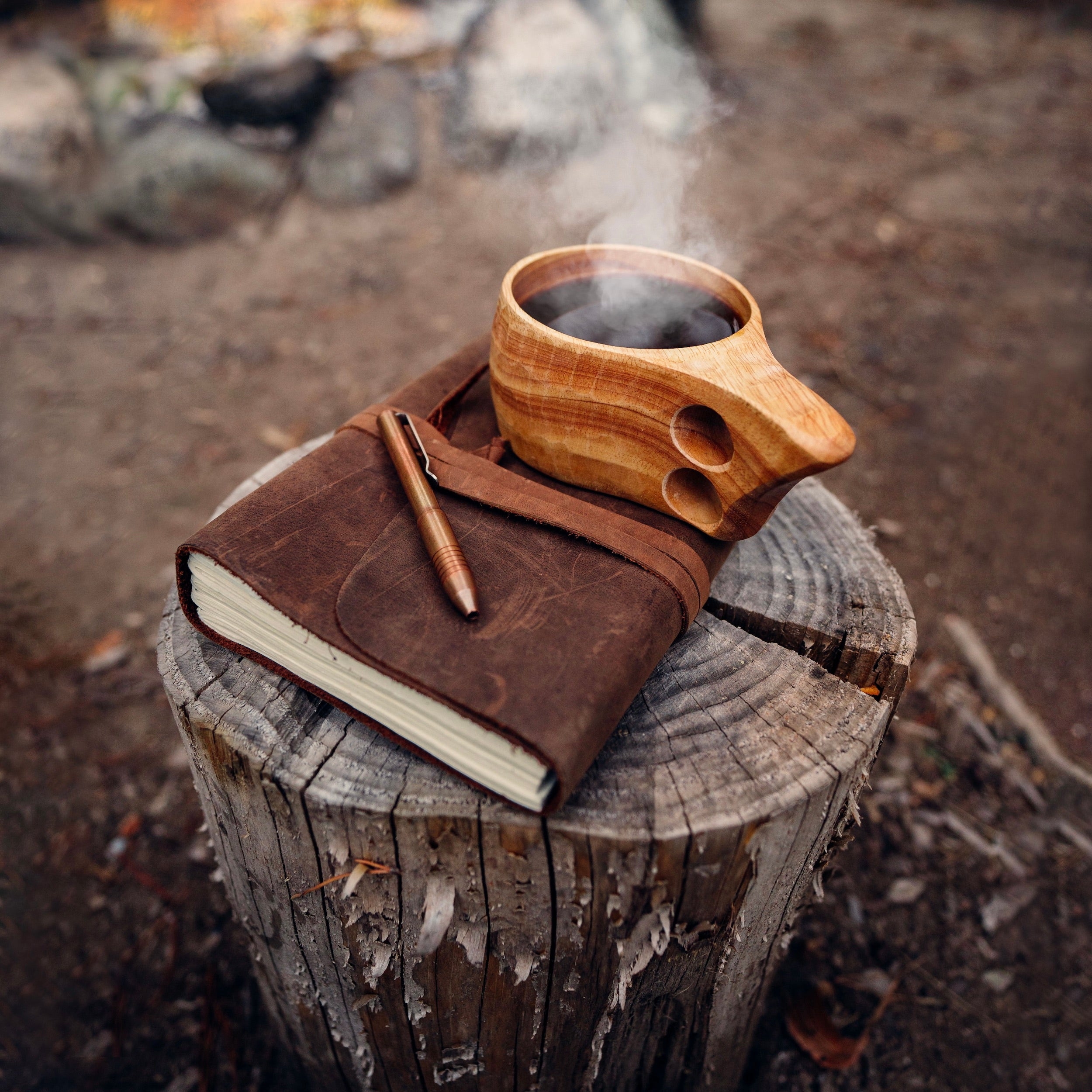 The Original Kuksa  camp cup from Überleben, filled with steaming hot coffee, sits atop a closed leather-bound journal. This setup sits on top of a tree stump against a backdrop of blurred rocks and nature.