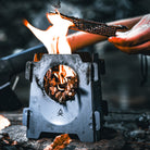 Close-up of the Überleben Stöker stove with a fire burning inside, fueled by twigs. A hand is adjusting a grill on top of the stainless steel stove. The image captures an outdoor setting with a blurred natural background, highlighting the compact metal structure of the Stöker stove.