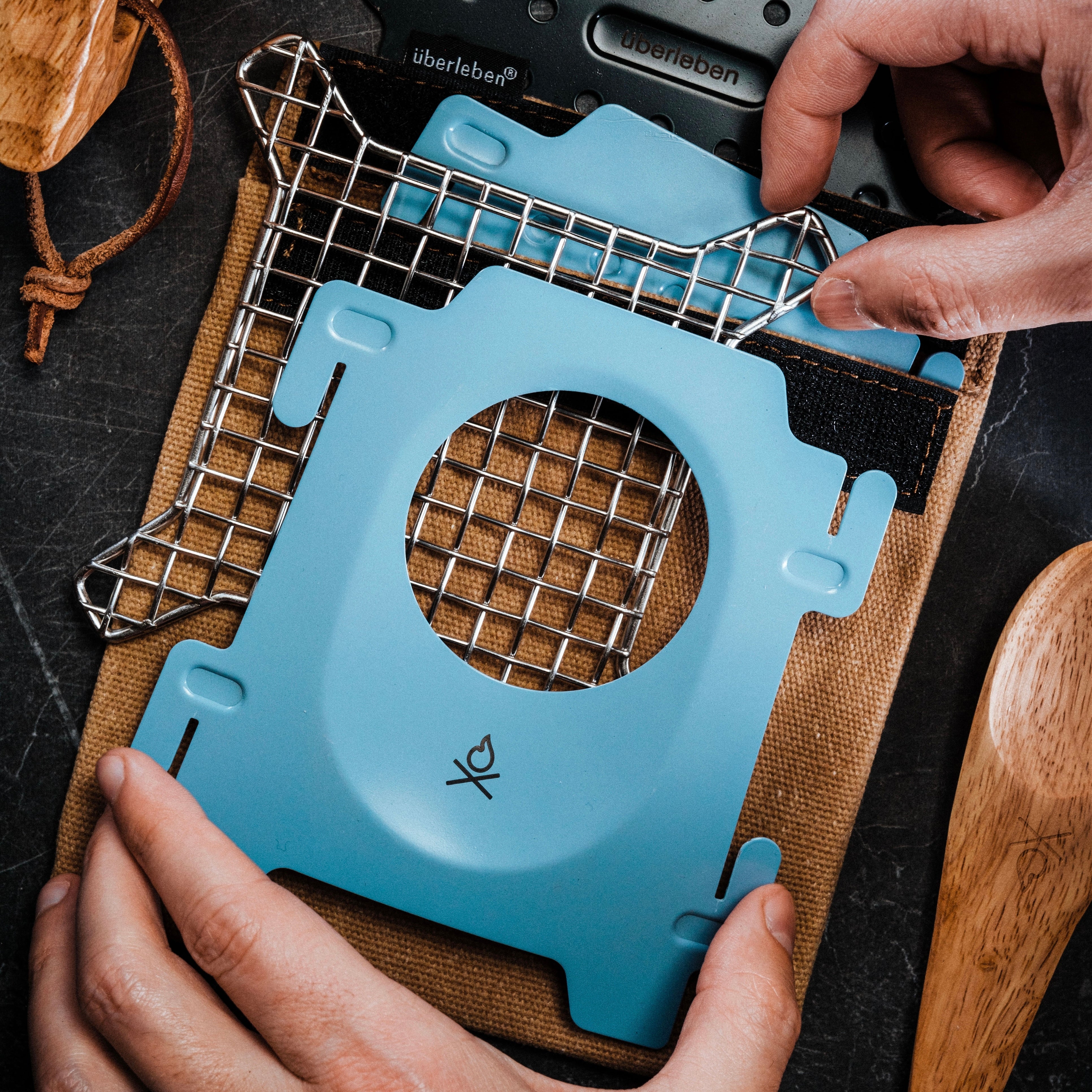 A pair of hands setting up the compact and portable bushcraft Stöker Stove by Überleben. The blue components of the stove and a stainless steel grid are being assembled over a waxed canvas pouch.