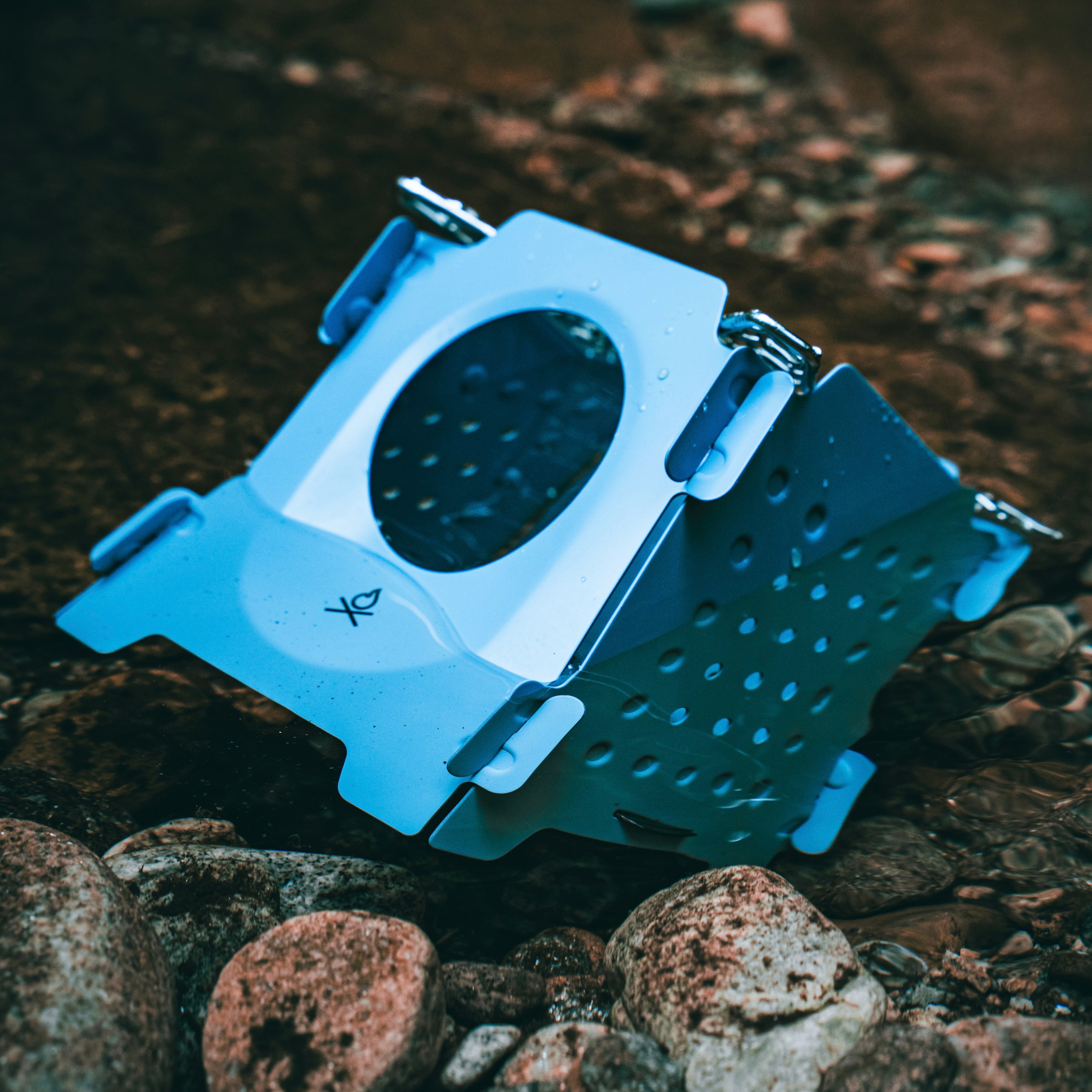 A close-up of the bushcraft Stöker Stove by Überleben, showcasing its blue stainless steel structure, resting on a bed of pebbles in a river. The stove features multiple foldable sections. It is tilted sideways, highlighting its perforated build.