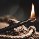 A lit fire starter, the Tindår  Wick & Bellow Kit. Branded "Überleben," its ultralight weatherproof paraffin-wax infused hemp wick burns steadily at one end, surrounded by dried leaves that suggest an outdoor setting.