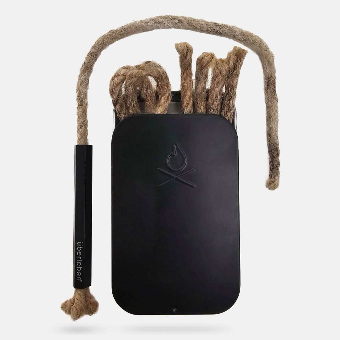 Three pieces of thick, paraffin-wax infused hemp wick from the Überleben Tindår Wick & Bellow Kit are arranged vertically in inside a black tin with a simple fire icon on the lid.