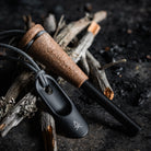 The Zünden 2.0 | Fire Starter, featuring a ferro rod with a wood composite handle engraved with the brand name "Überleben," rests on a pile of dried sticks. Beside it lies a black metal striker. This modern Flint & Steel setup appears on a dark, textured surface, ready for a fire-starting demonstration.