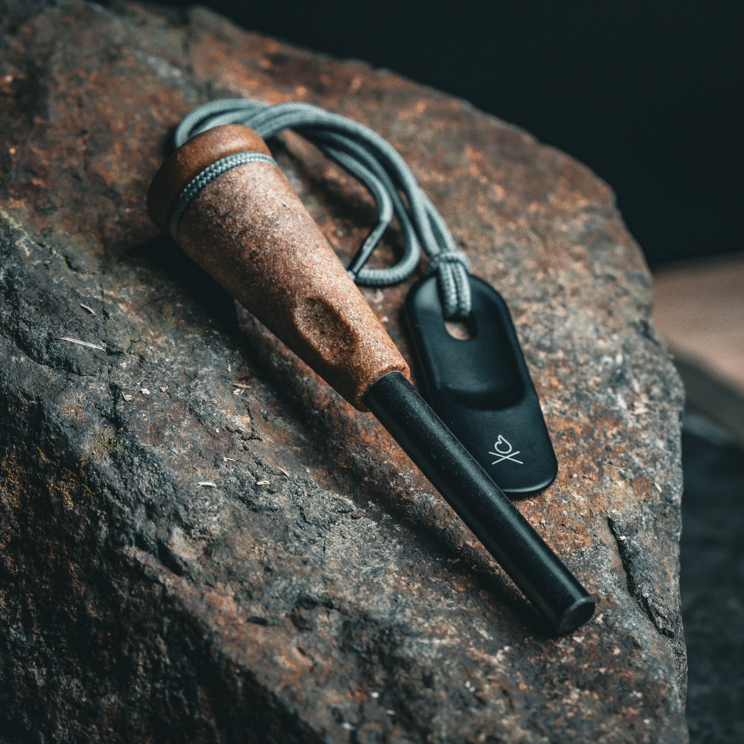 An Überleben fire starter, the  Zünden 2.0,  features a modern design, and includes a wood composite  handle and a black ferro rod that perfectly complements the rugged dark brown rock it is resting on. It also comes with a gray paracord loop attached to the fire starter, which is connected to a black metal striker showcasing a fire logo.