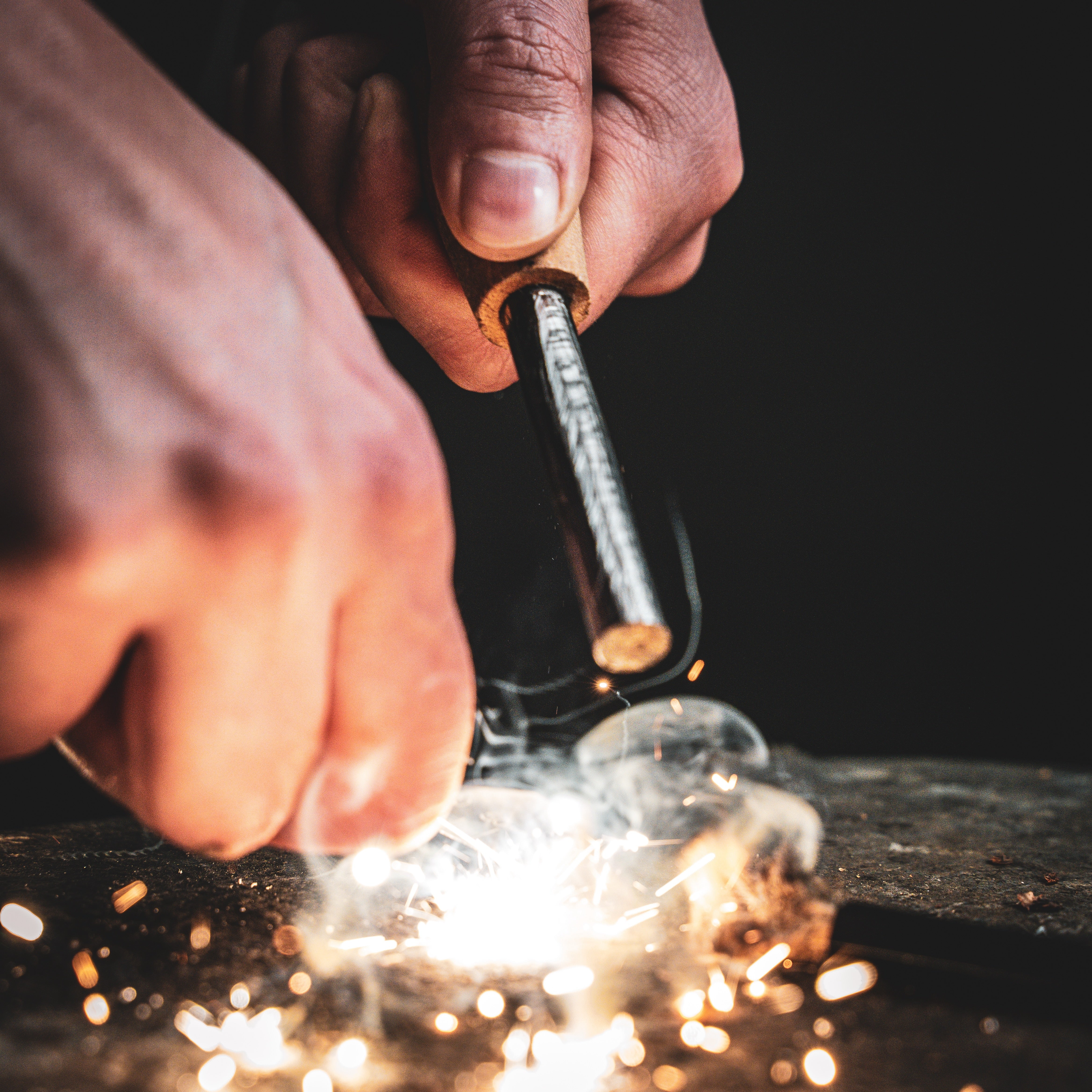 Close-up of hands striking a ferro rod, the Zünden | Fire Starter from Überleben, creating sparks against a dark background. The image captures the intensity and precision of the action, highlighting the glowing sparks flying out from the contact point, demonstrating the efficacy of Sanft-korr™ ferroceruim.