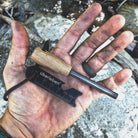 A hand with dirt-covered fingers holds the Überleben Zünden Fire Starter. The fire starter includes a ferrocerium rod with an Überleben wooden handle and a striker tool with measurement markings, both attached to a cord. The ferro rod is part of the durable Sånft-korr™ ferrocerium series.