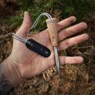 A hand holding an Überleben fire starter, the Zünden 2.0, which features a wood composite handle and a metal striker, alongside a gray lanyard. The background shows an outdoor, forest-like setting, perfect for this modern Flint & Steel.