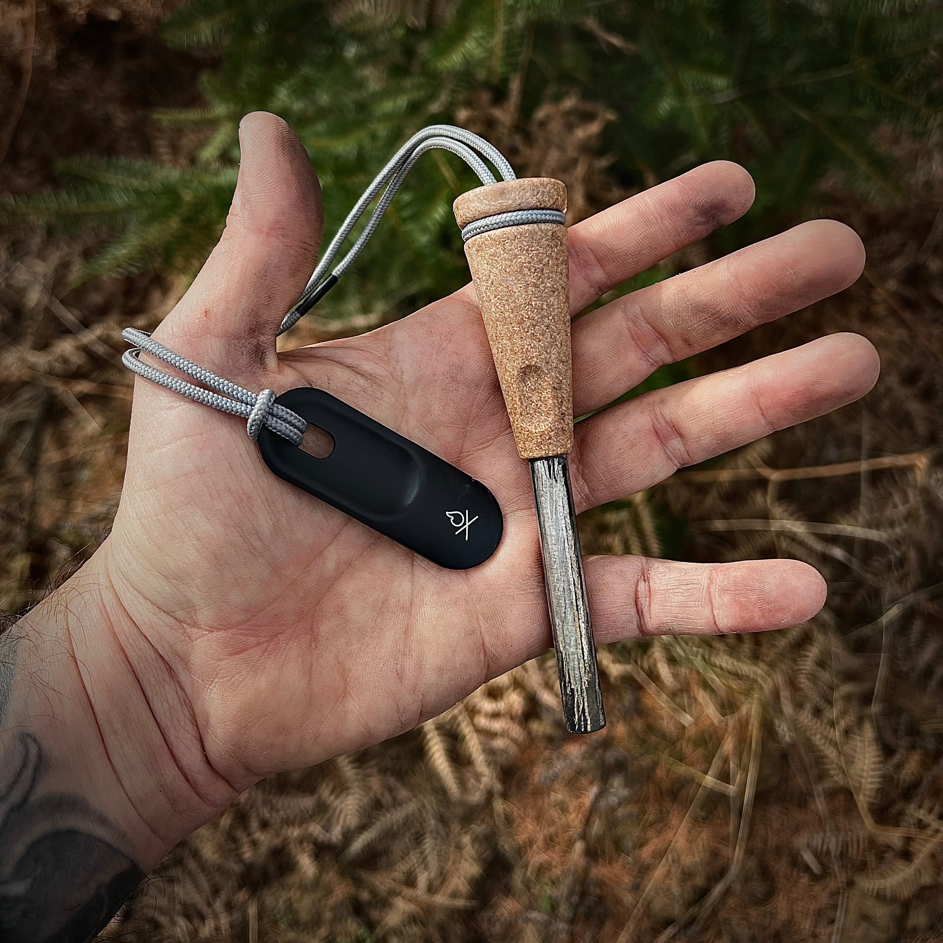 A hand holding the Überleben Zünden 2.0 Fire Starter, which features a wood composite handle and a metal striker, alongside a gray lanyard. The background shows an outdoor, forest-like setting, perfect for this modern Flint & Steel.