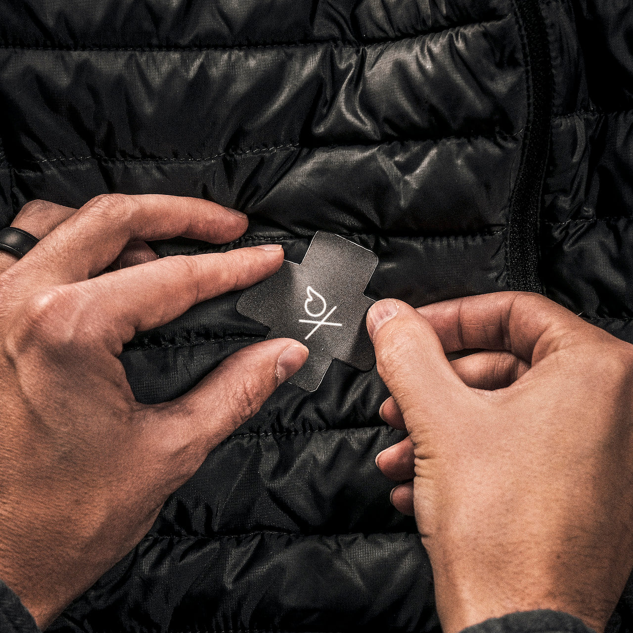 Two hands are seen patching a black puffy jacket with the Überleben x NOSO Fire Repair Patch in the shape of an X. The patch has a white fire icon near the center. The hands hold either end of the Peel & Stick Überleben repair patch, positioning it carefully over a tear in the jacket.