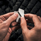 Close-up of two hands holding an Überleben x NOSO Fire Repair Patch, a small, X shaped repair patch. The background is a puffy jacket.