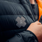 Close-up of a person adjusting a black winter jacket with an Überleben x NOSO | Fire Repair Patch in the shape of an X. The waterproof peel & stick patch, made by Überleben, features a simple fire icon.