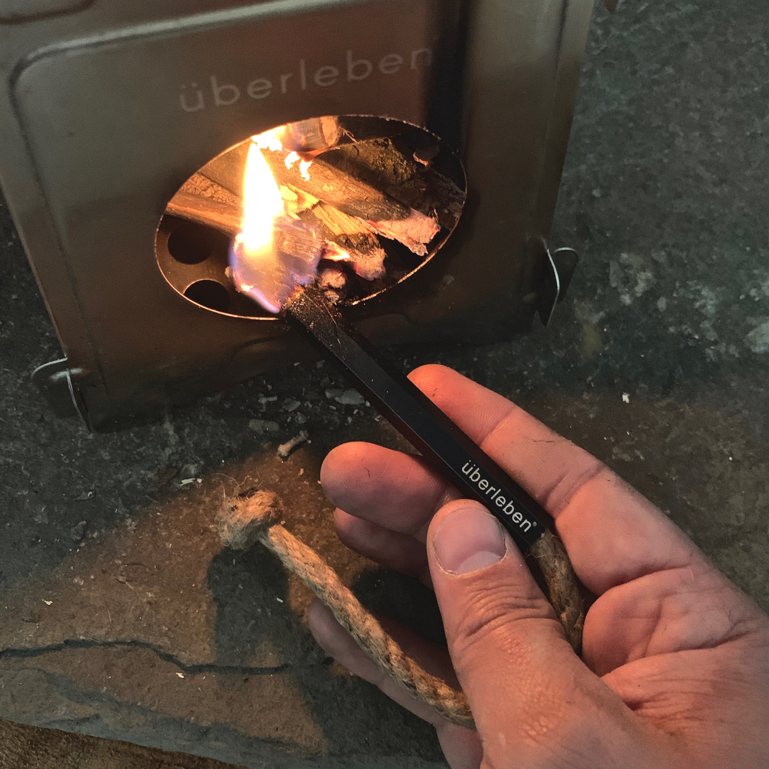 A hand holding an ultralight weatherproof Überleben Tindår Wick ignites kindling inside a small, twig burning stove. The Tindår Wick is a paraffin-wax infused hemp wick for an easy way to start your fire.