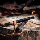 A lit fire starter, the Tindår Wick & Bellow Kit, rests on a stump with a ferro rod. Branded "Überleben," its ultralight weatherproof paraffin-wax infused hemp wick burns steadily at one end, surrounded by dried leaves that suggest an outdoor setting.