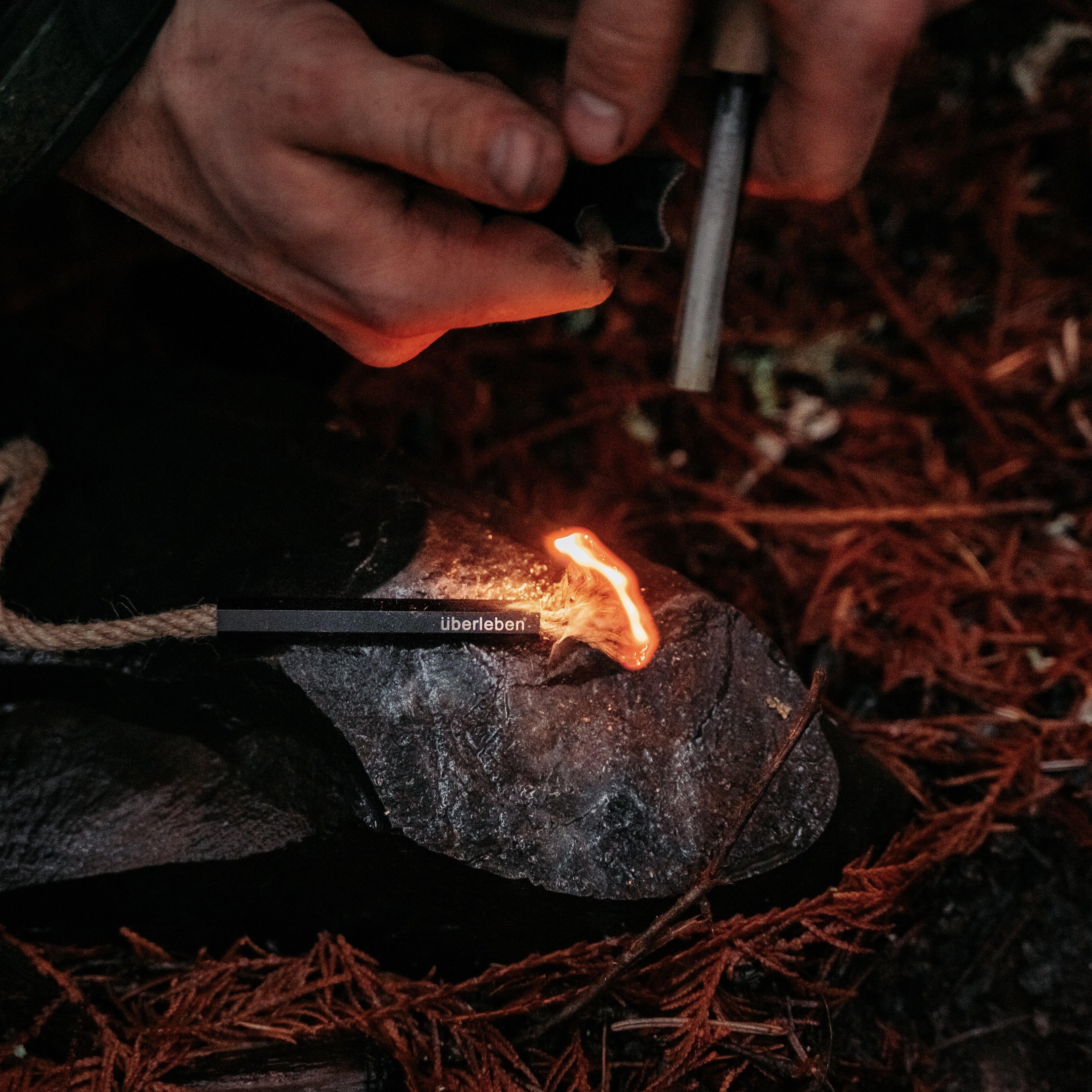 Close-up of a person using an Überleben ferro rod to ignite a small flame on a Tindår Wick & Bellow Kit, an ultralight weatherproof, paraffin-wax infused hemp wick. The ground is covered in brown pine needles. The person's hands are in focus, holding the tool and tinder.