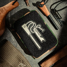 A flat lay of various survival gear items on a dark surface, featuring a camouflage-patterned pouch adorned with an Überleben Velcro Traddy Patch, showcasing stylized image of a Zünden Trad ferro rod, alongside a fire starter tool, and a green knit beanie.