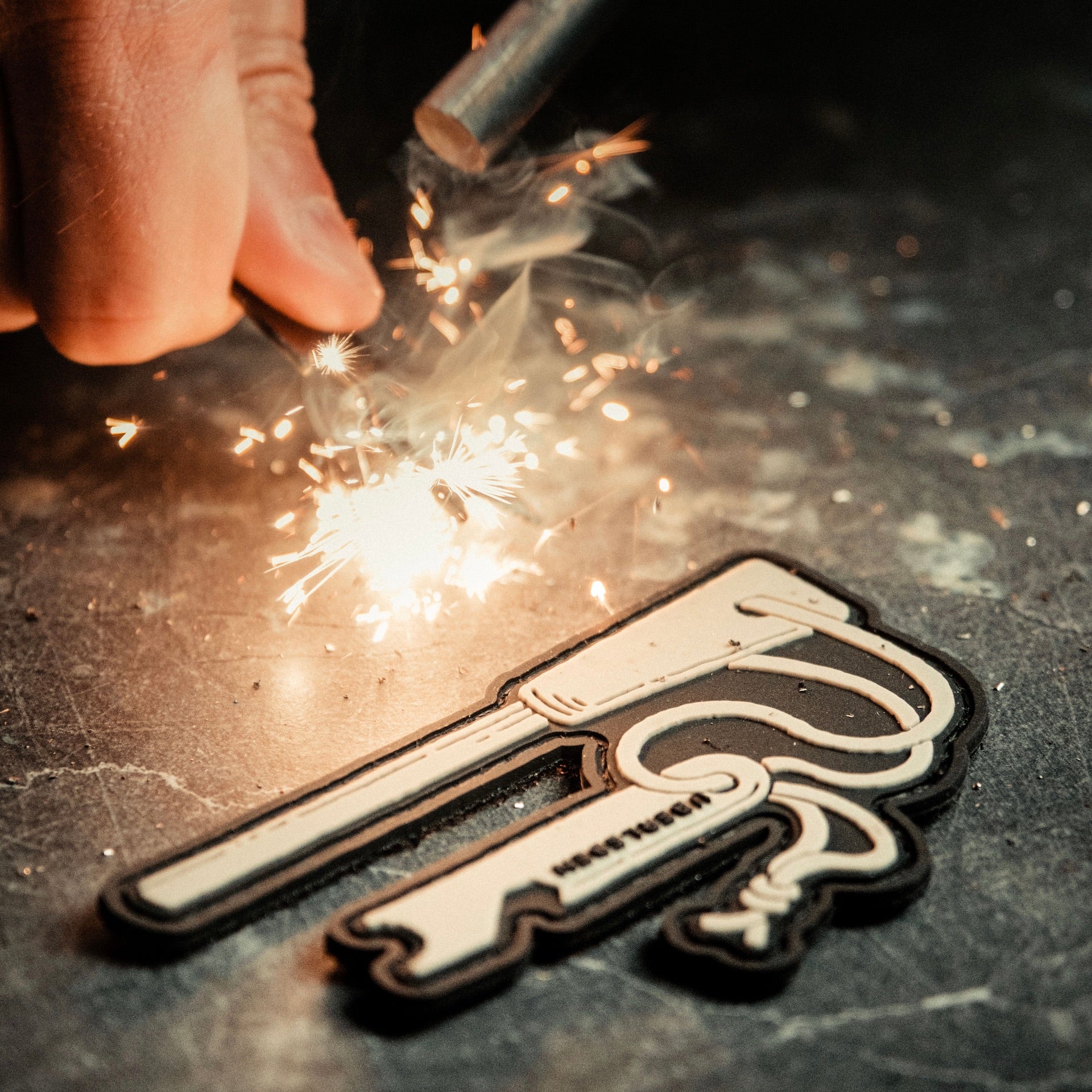 A close-up of a person's hand striking a ferro rod to produce sparks next to Überleben's Traddy Velcro Patch shaped like a Zünden fire starter. The patch features stylized design elements such as the wood handle and the striker.