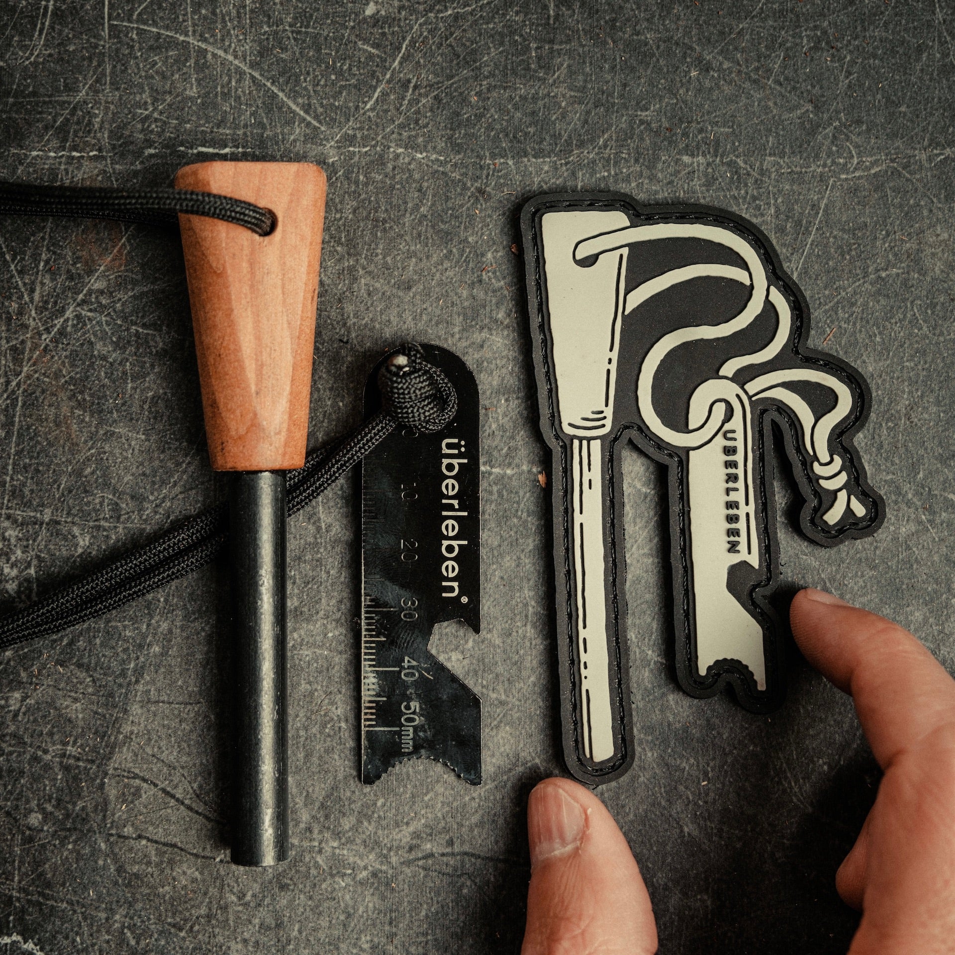 A hand holds a Traddy | Velcro Patch featuring a stylized ferro rod design and the text "Überleben." Next to it lies a Zünden fire starter, which is a wooden-handled ferro rod with a metal striker etched with the same brand's name. The background is a rustic, worn surface.