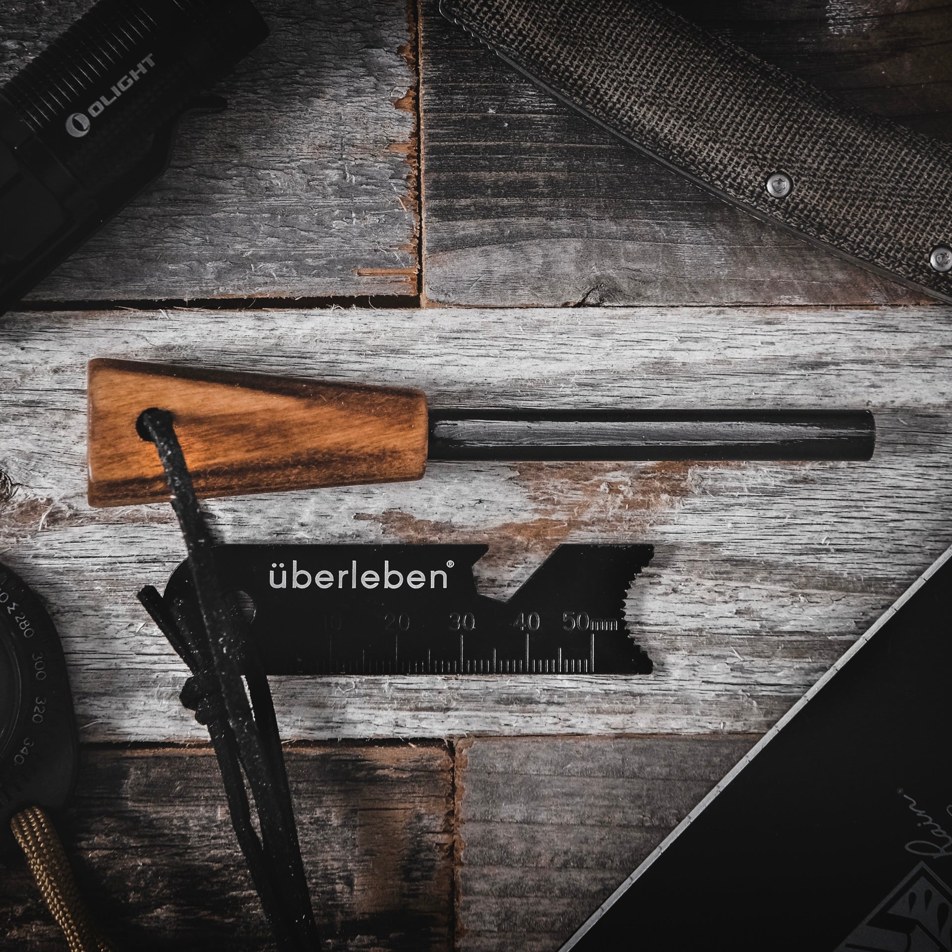 A flat lay photo displays a survival ferro rod on a weathered wooden surface. Items include the Zünden | Fire Starter, and a striker with the brand name "Überleben" on it. The scene emphasizes rugged, outdoor preparedness gear.